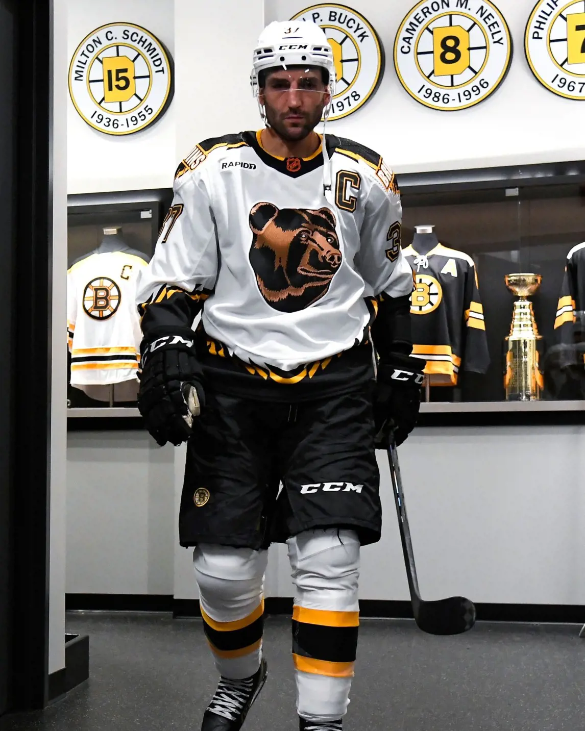 Patrice Bergeron wearing the Bruins mid 90s gold  jersey and carrying a hockey stick